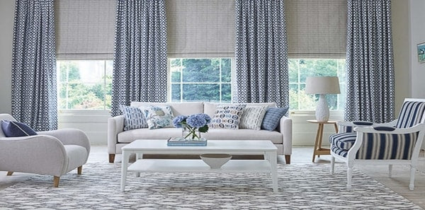 blue and white living room with baker lifestyle fabrics, blue geometric area rug, blue geometric drapery fabric, blue throw pillows, blue and white striped fabric, white coffee table, grey drapes, grey blue upholstered chair, white upholstered sofa