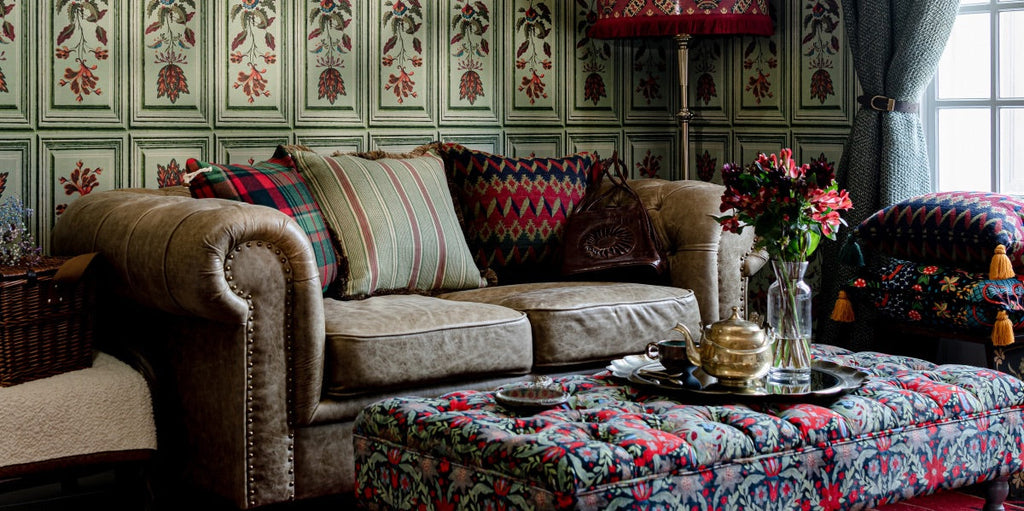 maximalist living room, maximalist design, paneled wallpaper, floral wallpaper, rich tones, upholstered coffee table, red and green throw pillows, metal floor lamp, suede upholstered sofa, silver serving tray, geometric fabric patterns, floral lumbar