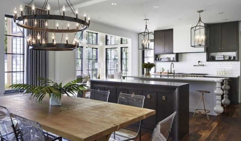 A Modern Country Kitchen in black and white by Charlie & Co in Elle Decor, tiered chandelier lighting, wooden dining room table, metal lantern pendants, dark grey drapery fabric, white vase, white island table, metal benches, clear dining room chairs