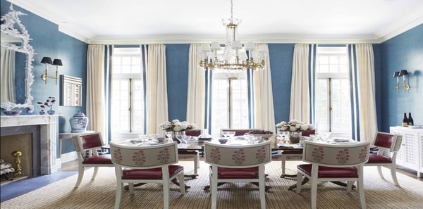 Blue dining room by Ashley Whitaker in Veranda Magazine, blue grasscloth, blue walls, tan area rug, red upholstered chair cushions, white decorative framed mirror, white and gold chandelier lighting, white draperies, blue wallpaper, textured walls