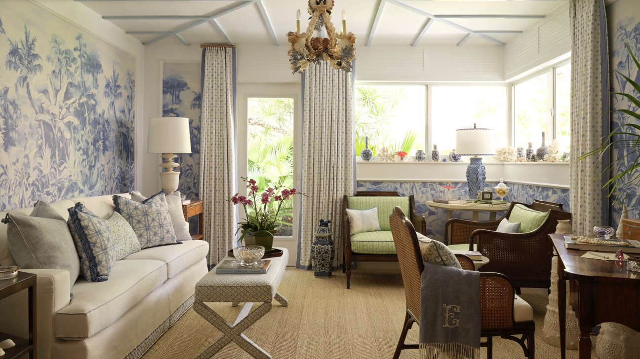 Bright family room with blue tropical wallpaper, neutral sofa, woven chairs, natural fiber rug, and large windows creating an airy, coastal-inspired space;  Brunschwig nouvelle wallpaper
