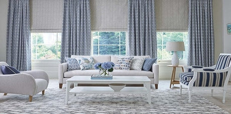 blue and white living room with baker lifestyle fabrics, blue geometric area rug, blue geometric drapery fabric, blue throw pillows, blue and white striped fabric, white coffee table, grey drapes, grey blue upholstered chair, white upholstered sofa