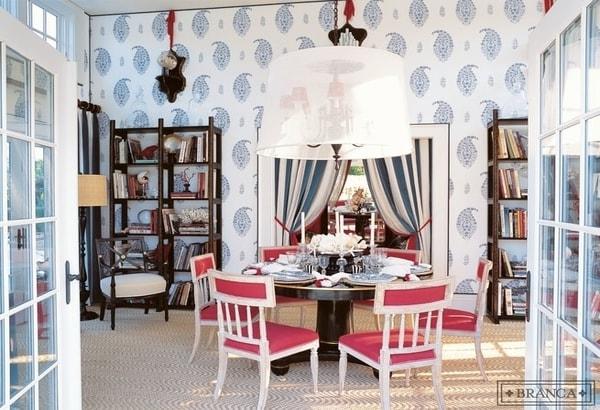Paisley dining room with Schumacher madras paisley wallpaper, paisley wallpaper, schumacher wallpaper, schumacher, pink upholstered dining chairs, tan textured area rugs, brown wooden bookshelves, white shaded pendant lighting, colorful design
