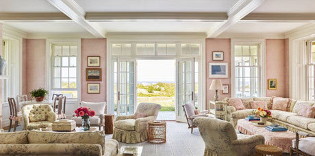 living room with pink walls, traditional coastal decor, beige and blue floral fabric, Miles Redd Block Island home, pink wallpaper, traditional upholstery, white upholstery fabric, geometric area rug, metal table lamp, rattan side tables, wall art