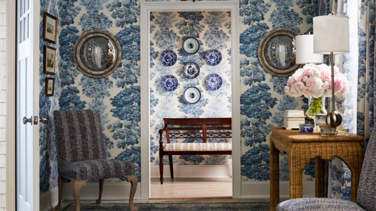 Elegant foyer with blue toile wallpaper, wicker console table, upholstered chairs, and decorative plate wall; Old World Weavers Central park wallpaper SCALA WWWP88583D-002 