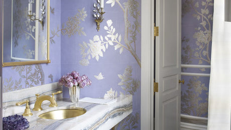 Lavender powder room with metallic floral wallpaper, marble vanity, gold sink and faucet, and brass sconces
