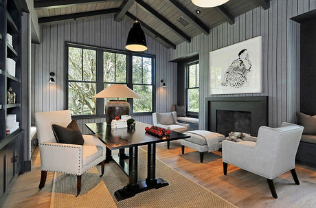 a rustic grey home office with striped grey wallpaper and light grey upholstered furniture. A large wood trestle desk anchors the space.