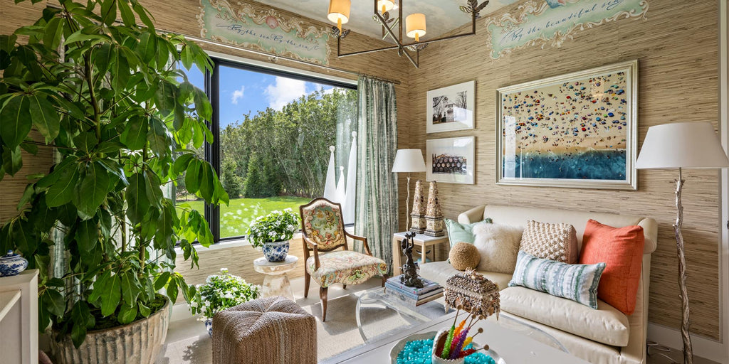 natural grasscloth walls, den, livingroom, barbara Ostrom, Holiday House Hamptons, grasscloth wallpaper, metal pendant lighting, gold accents, assorted colorful throw pillows, worldly design, upholstered ottoman, neutral area rug, orange and blue