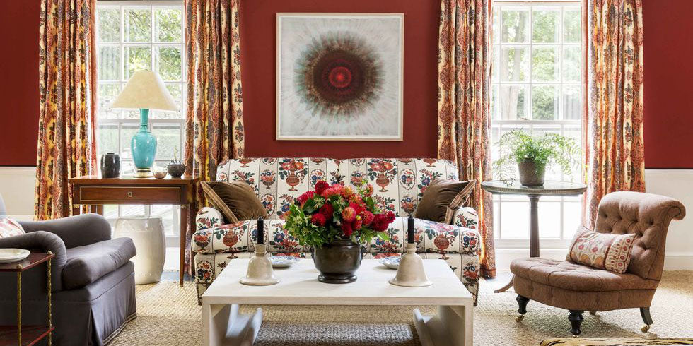 red walls, floral upholstered sofa, brown accent chairs, brown throw pillows, brown velvet, floral drapery, abstract wall art, white candlesticks, tan area rug, brown side table, wood accents, earth tone design, red and brown living room, blue lamp