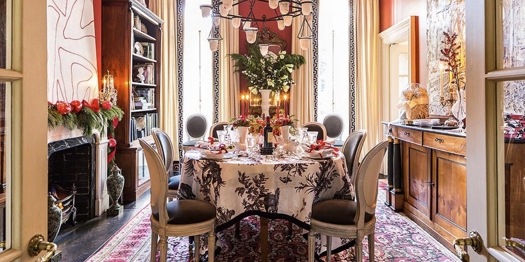 red walls, toile tablecloth, red traditional area rug, brown dining chairs, black and white chandelier lighting, abstract geometric accent wallpaper, brown and white tablecloth, wood accent decor, wood bookshelves, white vase, white black draperies