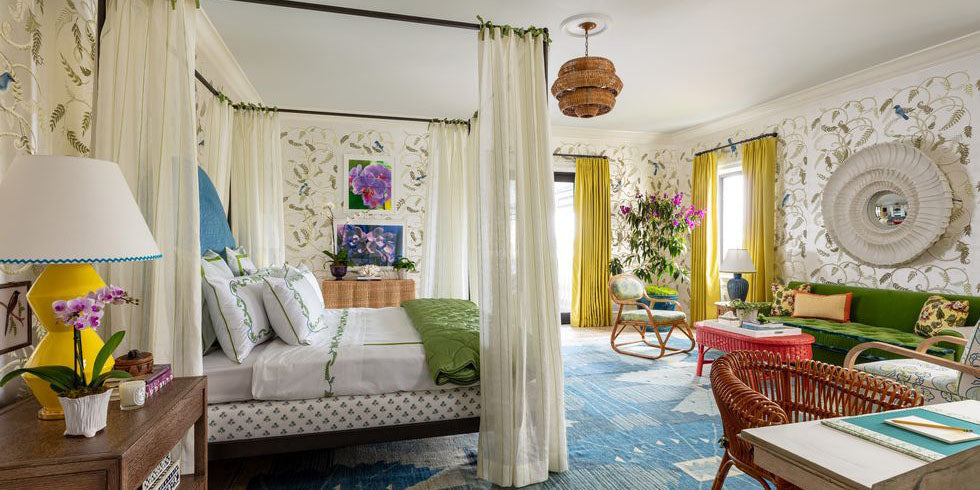 bedroom with floral and bird wallpaper, katie ridder kips bay palm beach showhouse, canopy bed, four poster bed, blue abstract area rug, yellow drapery, white canopy, white desk, yellow table lamp, large framed decorative mirror, wall art, green sofa