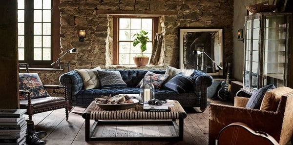 Ralph Lauren home decor in a modern country setting with a blue sofa and stone walls, blue velvet upholstery, blue and tan pillows, stone walls, rustic design, rustic coffee table, brown upholstered chair, wood floors, wood hutch, arm floor lamps