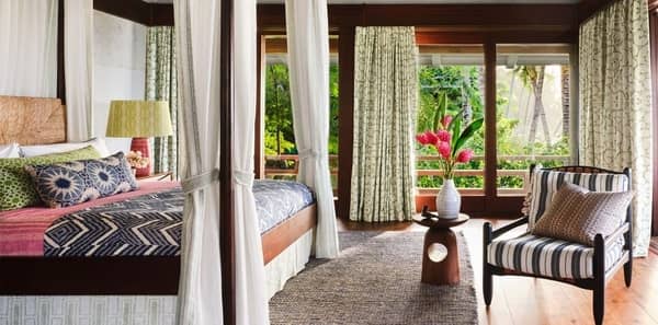 Tropical bedroom, Elle Decor Martyn Lawrence Bullard, green floral drapery, chevron coverlet, canopy bed, green drapery, grey area rug, blue striped upholstered chair, wood accent decor, white walls, white vase, brown four poster bed, blue pillows