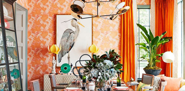 colorful design, orange and pink abstract wallpaper, heron wall art, white geometric hutch, blue upholstered dining chairs, gold abstract chandelier lighting, white floor lamp, white statuettes, glass dining room table, blue trim, large blue vase