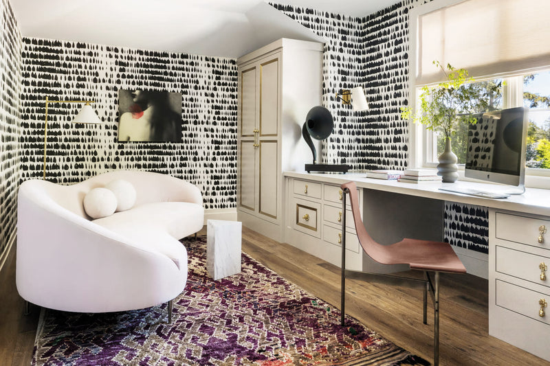 Modern home office with black and white patterned wallpaper, built-in desk, neutral cabinetry, sculptural chair, and warm wood flooring; Schumacher QUEEN OF SPAIN wallpaper, white cultural sofa