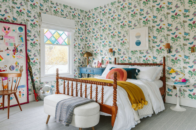 Kids bedroom with butterfly and floral wallpaper, wooden bed, colorful accents, white bedding, and bright natural light