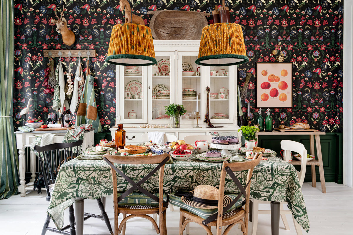 Dining room with MINDTHEGAP folk floral wallpaper, a green printed tablecloth and amber pendant lamps.